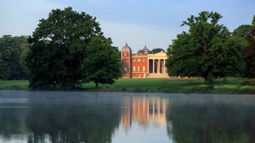 Neoclassical red-brick mansion with a columned portico, viewed across a calm lake with soft mist, reflecting the building and surrounding trees in the water.
